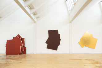 A large gallery space with wood floors, a white wall and verical skylights with 3 large paintings. The leftmost is comprised of multiple layers of a jagged shape in a rust color. In the center and on the right are angular shapes in brown and gold, respectively.